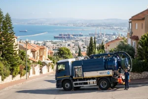 ביובית של החברה חונה בבטחה ברחוב משופע בחיפה, עם נוף המפרץ ברקע.